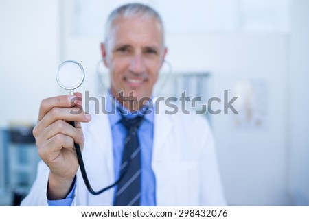 Happy doctor smiling at camera and showing his stethoscope in medical office