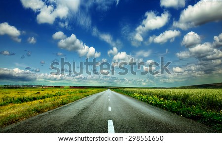 image of wide open prairie with a paved highway stretching out as far as the eye can see with beautiful small green hills under a bright blue sky in the summer time Royalty-Free Stock Photo #298551650