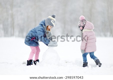 Two funny adorable little sisters making a snowman together in beautiful winter park during snowfall