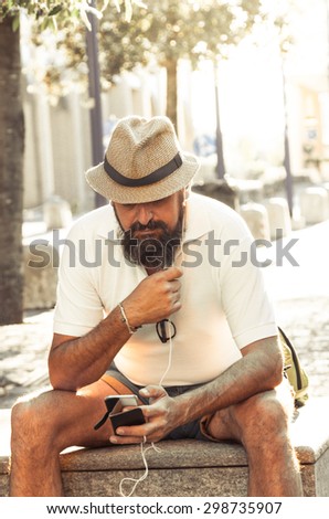 Urban Technology concept. Stylish adult hipster with long beard and mustaches wearing a straw hat is listening music with earphones outdoor at sunset. Sun flare digitally enhanced. 