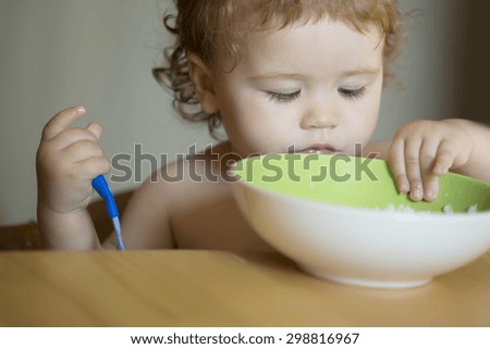 Portrait of curious small pretty baby boy with blonde curly hair and round cheecks eating from green plate with hand holding spoon closeup, horizontal picture