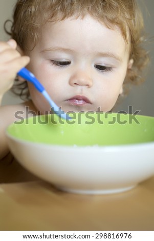 Portrait of interesting little male kid with blonde curly hair and round cheecks eating from green plate with spoon closeup, vertical picture