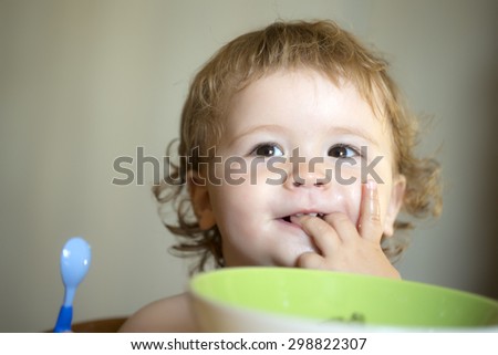 Portrait of sweet small smiling baby boy with blonde curly hair and round cheecks eating from green plate holding spoon and lick fingers closeup, horizontal picture