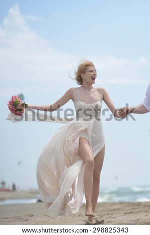 Happy beautiful young laughing bride in beige dress holding wedding bouquet of coral roses running on sea beach coast on blue sky background, vertical picture