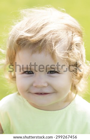 Portrait of small smiling beautiful male child with blonde curly hair looking forward outdoor sunny day on natural green background, vertical picture
