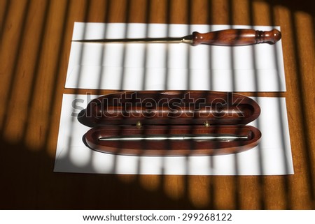 Metallic grey ball-point pen in brown wooden open case and sharp golden paper knife lying on two white envelopes on office table on jalousie shadow background, horizontal picture