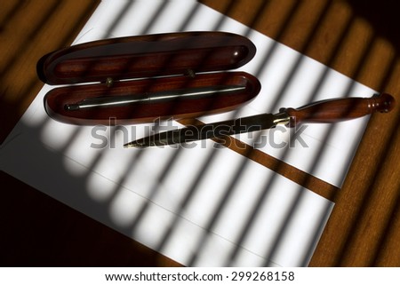 Silver grey biro pen in brown wooden open case and sharp golden paper knife lying on two white envelopes on office table on jalousie shadow background, horizontal picture
