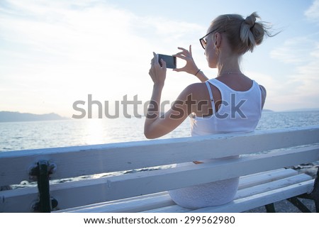 Beautiful young blonde woman using mobile phone, sitting on bench. Summer photo. Coastal view.