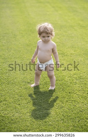 Little pretty curious male child with blond curly hair in white nappy standing making shadow on field with fresh green grass sunny day outdoor in summer, vertical picture