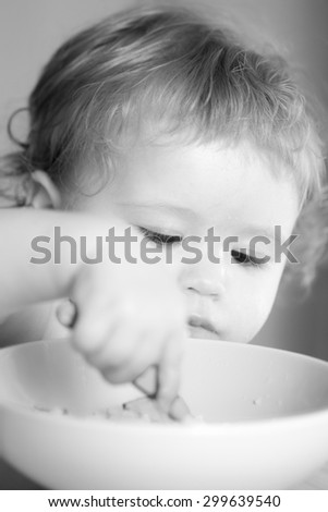 Portrait of serious small cute male kid with curly hair and round cheecks eating from plate with spoon black and white closeup, vertical picture