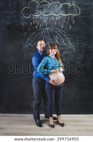 Conceptual shot of husband hugging pregnant woman under umbrella at rain against chalk drawing