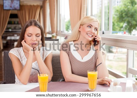 Cheerful girls are sitting at the table in cafe and drinking juice. The blond girl is talking on the phone and smiling. Her friend is bored. She is looking down sadly and leaning her head on hands