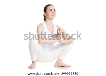 Sporty smiling beautiful young woman in white sportswear sitting in squat, Garland Pose, Malasana with hands in Namaste, studio full length shot on white background, three-quarters view, isolated