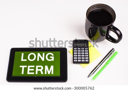 Business Term / Business Phrase on Tablet PC - Cup of coffee, Pens, Calculator and a green/yellow note pad on a White surface - White Word(s) on a green background - Long Term
