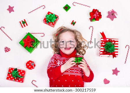 Child opening Christmas presents. Little girl in knitted winter sweater with present box. Kids open gifts. Toddler kid on the floor under decorated Xmas tree. Children play with gift box and candy.