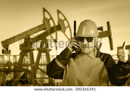 Adult industrial worker in glasses, with helmet camera, talking on radio transceiver, on a  oil field background. Toned sepia. 