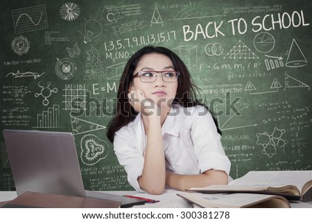 Portrait of a female high school student wearing glasses in class and looks pensive with laptop and books on the desk