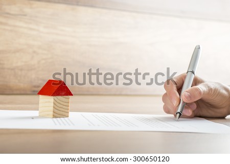Closeup of new homeowner signing a contract of house sale or mortgage papers with a wooden toy house on the document. Suitable for real estate concept. 