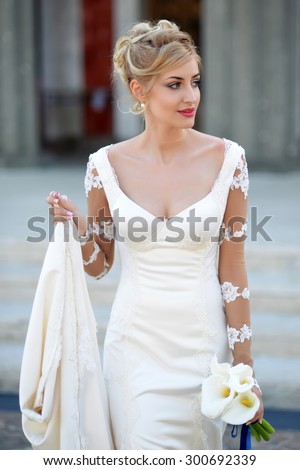 Beautiful young blonde fiancee with elegant hairstyle in white wedding dress with long train holding bouquet of calla flowers standing outdoor on stone building background, vertical picture