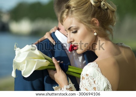 Lovely young wedding couple of guy in blue jacket embracing blonde woman in white dress with red lips holding bunch of calla flowers standing on sunny outdoor background, horizontal picture 