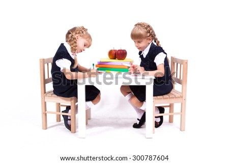 Two cute curious girl twins on white background sitting at a table surrounded by books in elegant dresses and painted. The picture with depth of field
