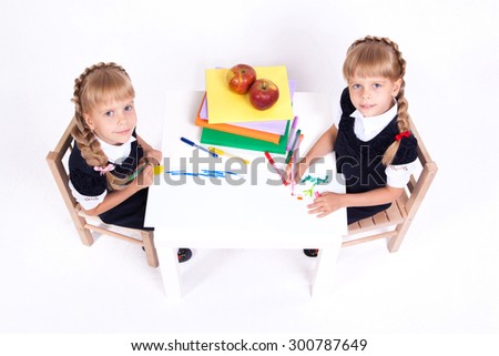 Two cute curious girl twins on white background sitting at a table surrounded by books in elegant dresses and painted. The picture with depth of field