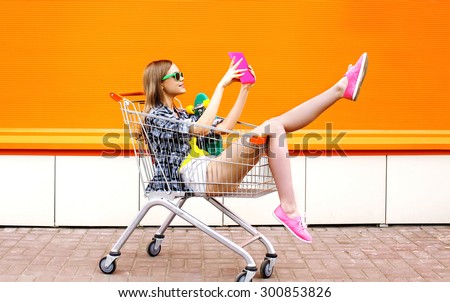 Fashion smiling hipster woman having fun taking picture selfie on the digital tablet pc wearing a sunglasses with skateboard sitting in the shopping trolley cart outdoors