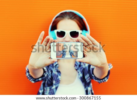 Pretty cool girl makes self-portrait on the smartphone and listens to music in headphones against colorful orange wall, screen phone closeup
