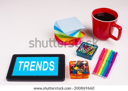Business Term / Business Phrase on Tablet PC - Colorful Rainbow Colors, Cup, Notepad, Pens, Paper Clips, White surface - White Word(s) on a cyan background - Trends