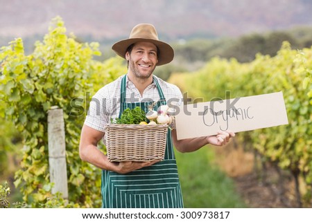 Young happy farmer holding a basket of vegetables and a sign in the field