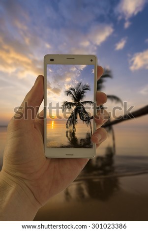 Tourist hand holding smart phone, taking photo of Thailand beach sunset palm tree