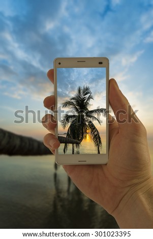 Tourist hand holding smart phone, taking photo of Thailand beach sunset palm tree