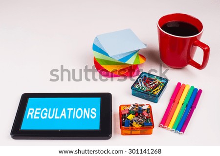 Business Term / Business Phrase on Tablet PC - Colorful Rainbow Colors, Cup, Notepad, Pens, Paper Clips, White surface - White Word(s) on a cyan background - Regulations