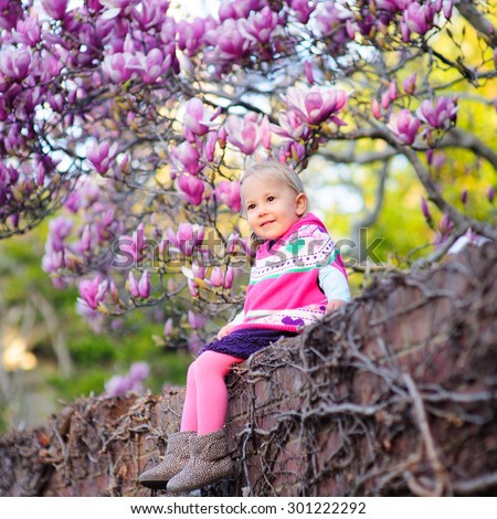 Outdoor portrait of an adorable happy little toddler girl wearing warm knitted poncho, tights and boots enjoying sunshine on a beautiful spring day in the garden with magnolia trees in full bloom