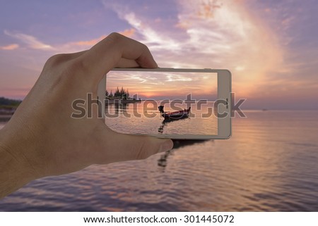 Tourist hand holding smart phone, taking photo of Thailand beach sunset boat