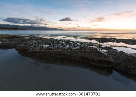Long exposure of a beach with smooth waters on the north shore of Oahu, Hawaii during dusk. Tide pools in the foreground and the western tip of the island on the horizon. 
