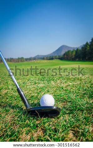 chipping a golf ball onto the green with driver golf club. Green grass with forrest and mountains in the background. Soft focus or shallow depth of field. Back view
