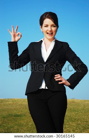 Beautiful young businesswoman outdoors in a field with a blue sky giving the okay sign.
