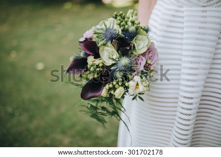 bride in a white dress holding a bouquet of purple flowers and greenery on the background of green grass