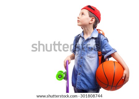 Pensive school boy holding a skateboard and a basketball on white background