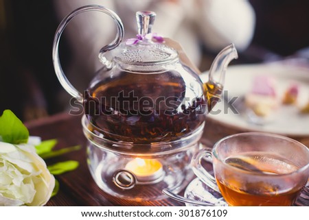 Beautiful warm picture of transparent teapot kettle with tasty green black tea with apple, lemon and ginger on a table with candles and with dessert in the background