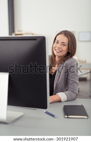 Pretty Young Office Woman Sitting at her Desk with Computer and Notes, Looking at the Camera with a Smiling Face.