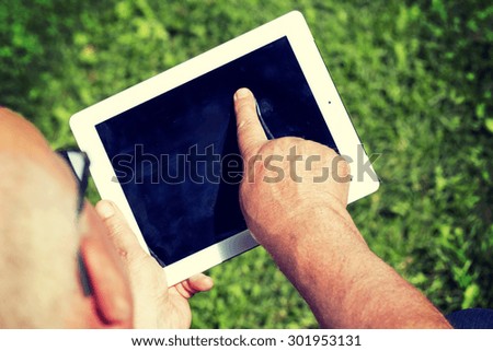 An elderly man is browsing the internet with a tablet outdoor with a green grass on the background. Image has a strong vintage effect applied.