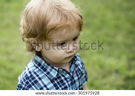 Portrait of small curious boy child with blonde curly hair in chequered blue shirt standing on green grass lawn looking away on natural background outdoor closeup, horizontal picture