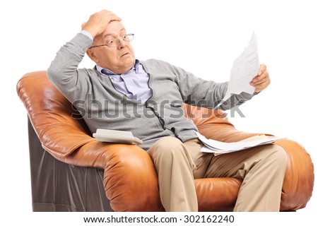 Shocked senior gentleman looking at his bills in disbelief seated in an armchair isolated on white background Royalty-Free Stock Photo #302162240