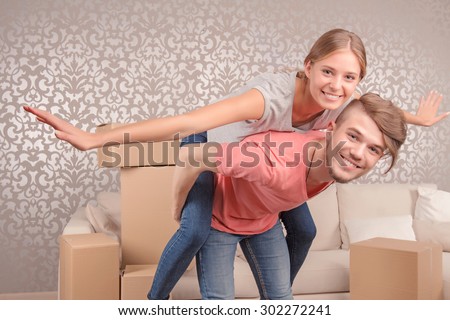 I can fly.  Delighted smiling young husband bending and holding his beautiful wife on his back while having fun in their new flat
