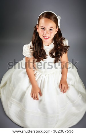 Beautiful young girl dressed in white. First Communion. Perfect teeth and smile, long curly hair, hands folded. Dark background, studio shoot.