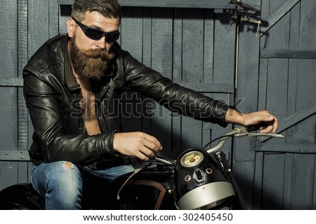 Handsome brutal unshaven male biker with long beard in brown leather jacket jeans and sun glasses sitting in garage on motorcycle looking forward on grey wooden background, horizontal picture