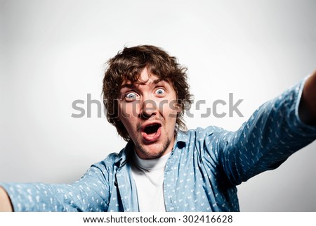 Close up portrait of a young shocked man holding a smartphone digital camera with his hands and taking a selfie self portrait of himself standing against grey background