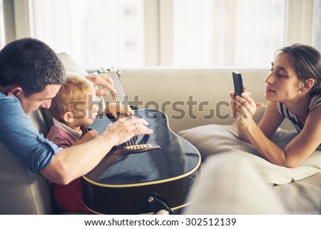 Family around guitar, young child is trying to play it, mom is taking photo 